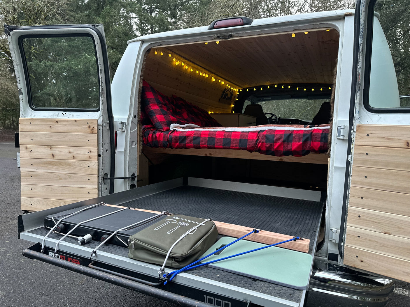 This cozy RV rental camper van features a warm plaid bed, string lights, and a pull-out kitchen for outdoor cooking.