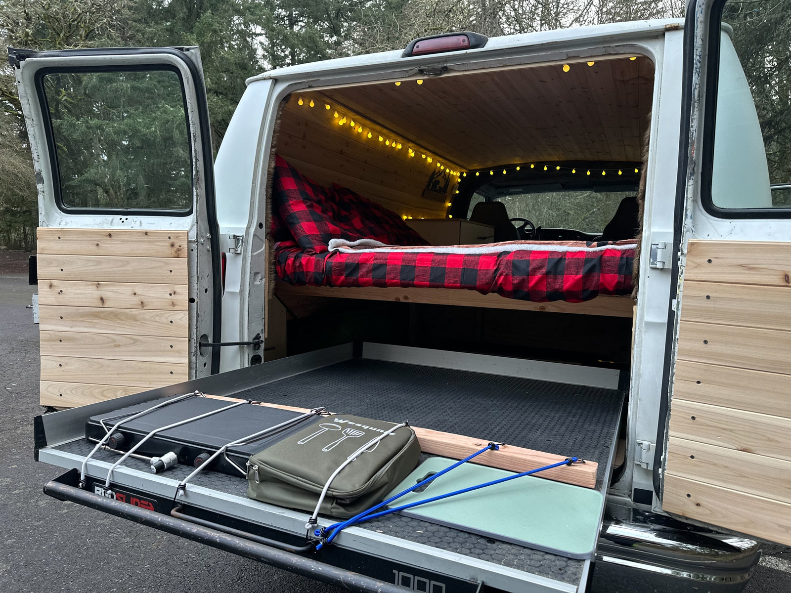 This cozy RV rental camper van features a warm plaid bed, string lights, and a pull-out kitchen for outdoor cooking.
