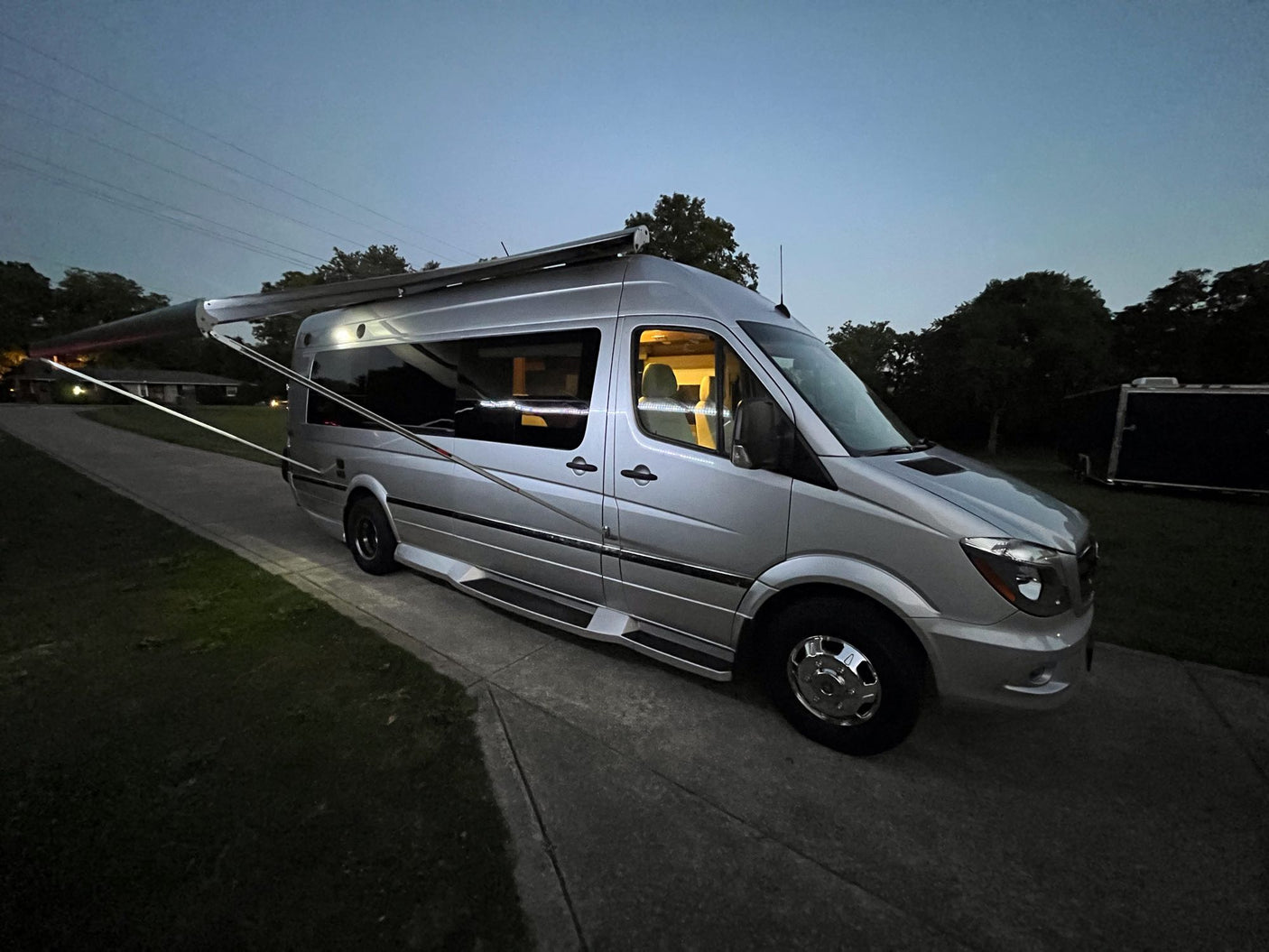 Experience van life! This silver Sprinter RV rental features an extended awning & illuminated interior at dusk.|Front view of a silver Mercedes-Benz Sprinter RV rental