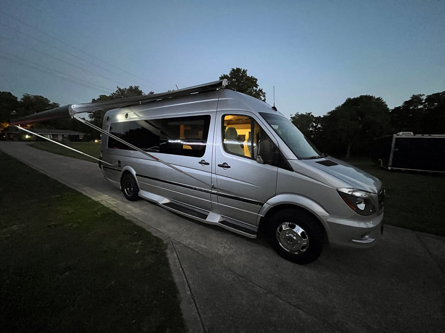 Experience van life! This silver Sprinter RV rental features an extended awning & illuminated interior at dusk.|Front view of a silver Mercedes-Benz Sprinter RV rental