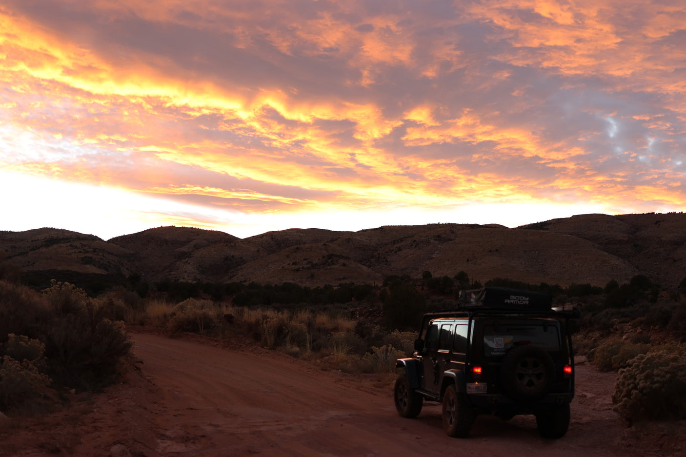 A dark RV rental Jeep with rooftop tent on a dirt road, facing dark mountains under a vibrant orange sunset sky.