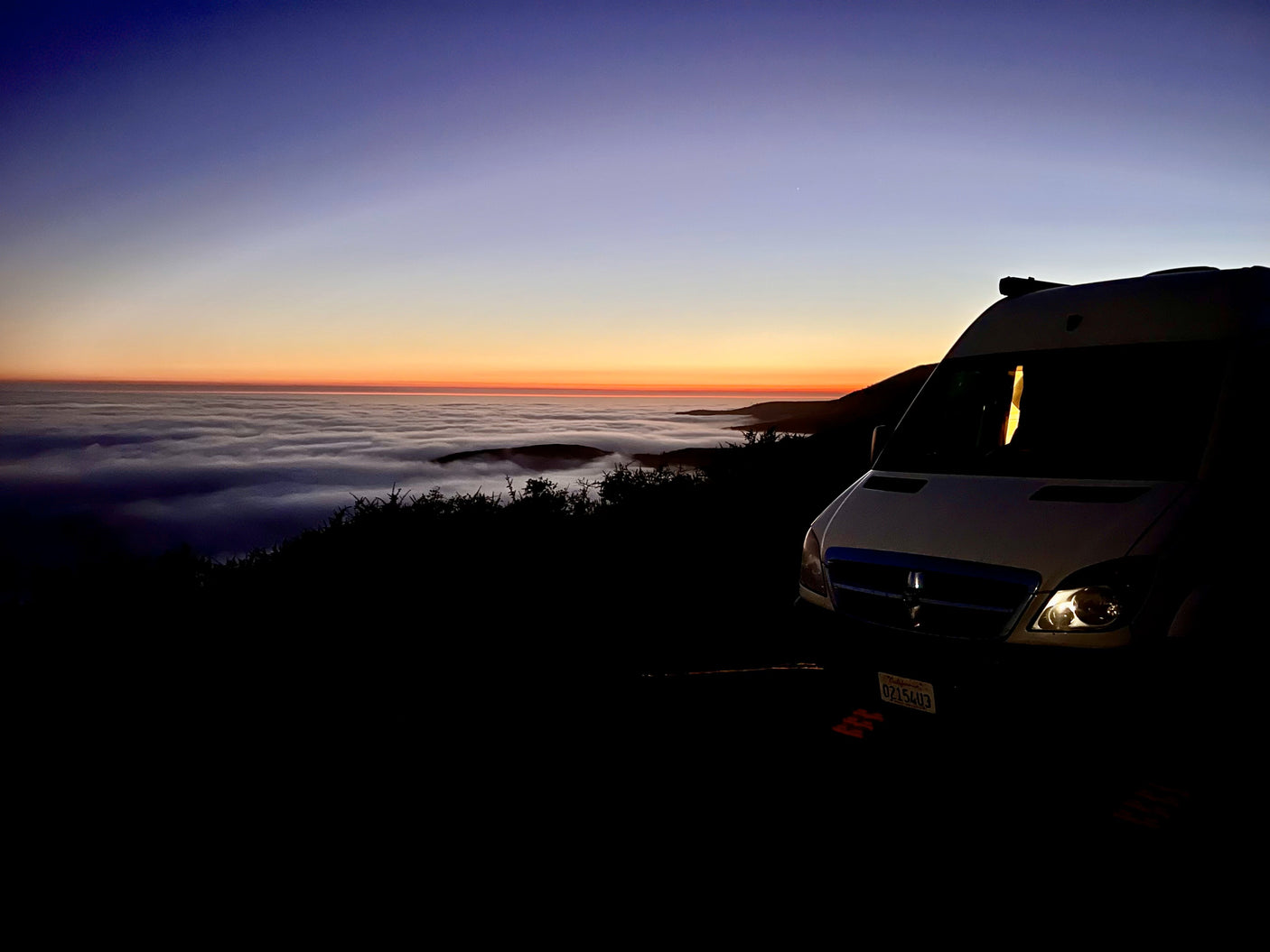 An RV rental parked at dawn, overlooking a sea of clouds and a vibrant blue-to-orange sky. Scenic mountain adventure awaits.