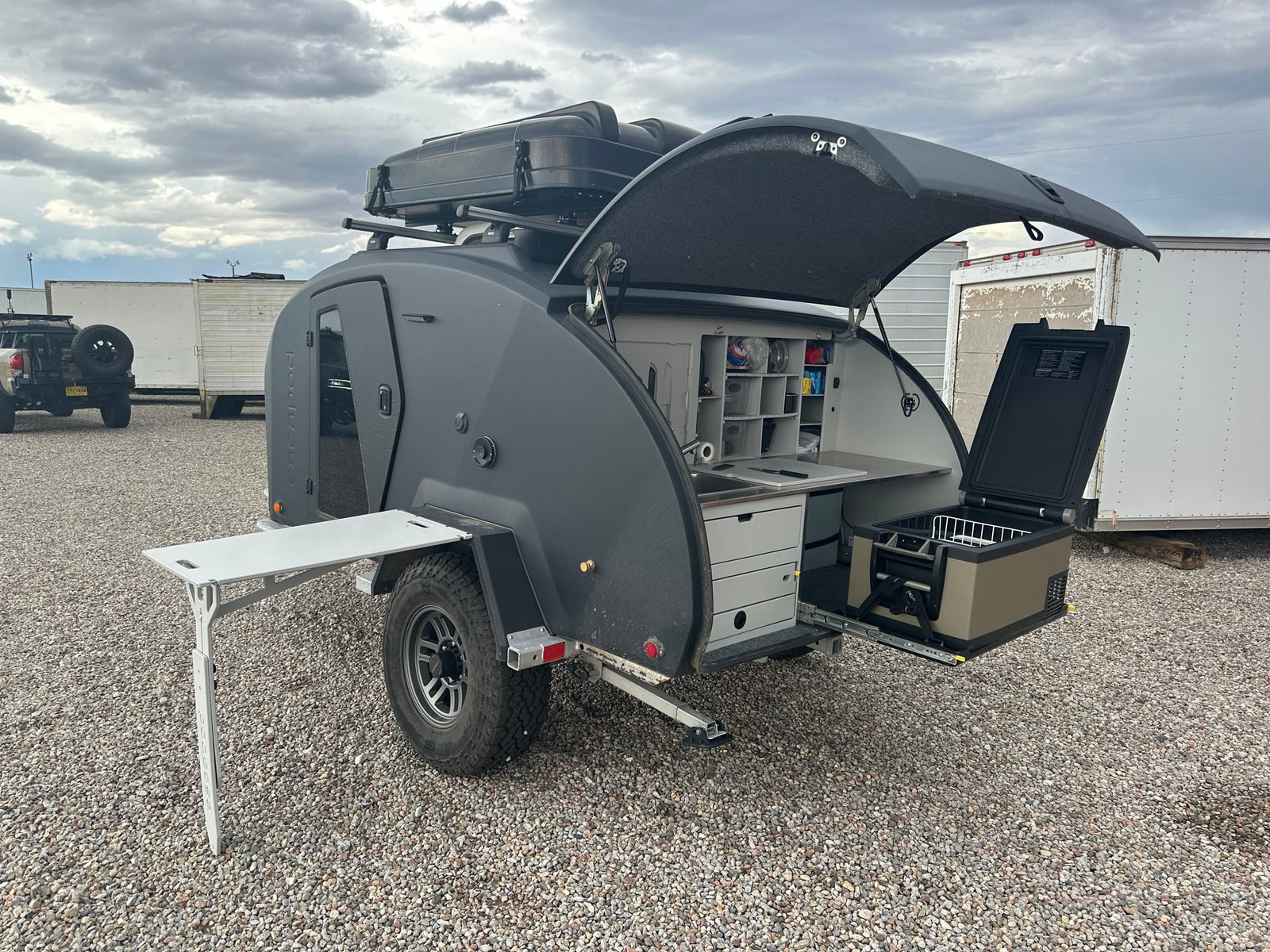 A sleek dark gray RV rental teardrop trailer with its rear hatch open, revealing a fully equipped outdoor kitchen and pull-out fridge.