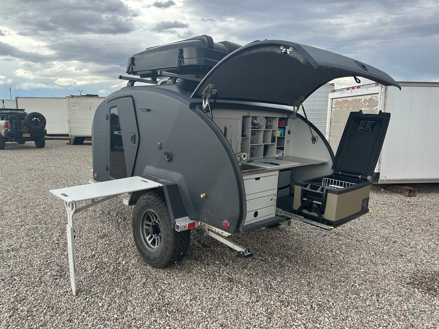 A sleek dark gray RV rental teardrop trailer with its rear hatch open, revealing a fully equipped outdoor kitchen and pull-out fridge.