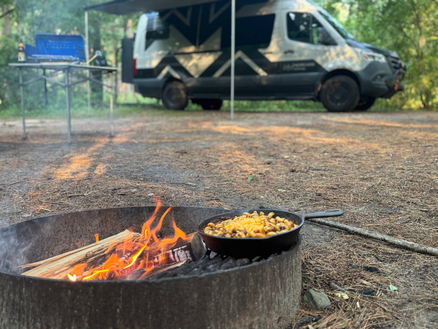 RV rental evening: a skillet of chili mac cooks over a campfire, with a camper van and outdoor kitchen setup in the forest.