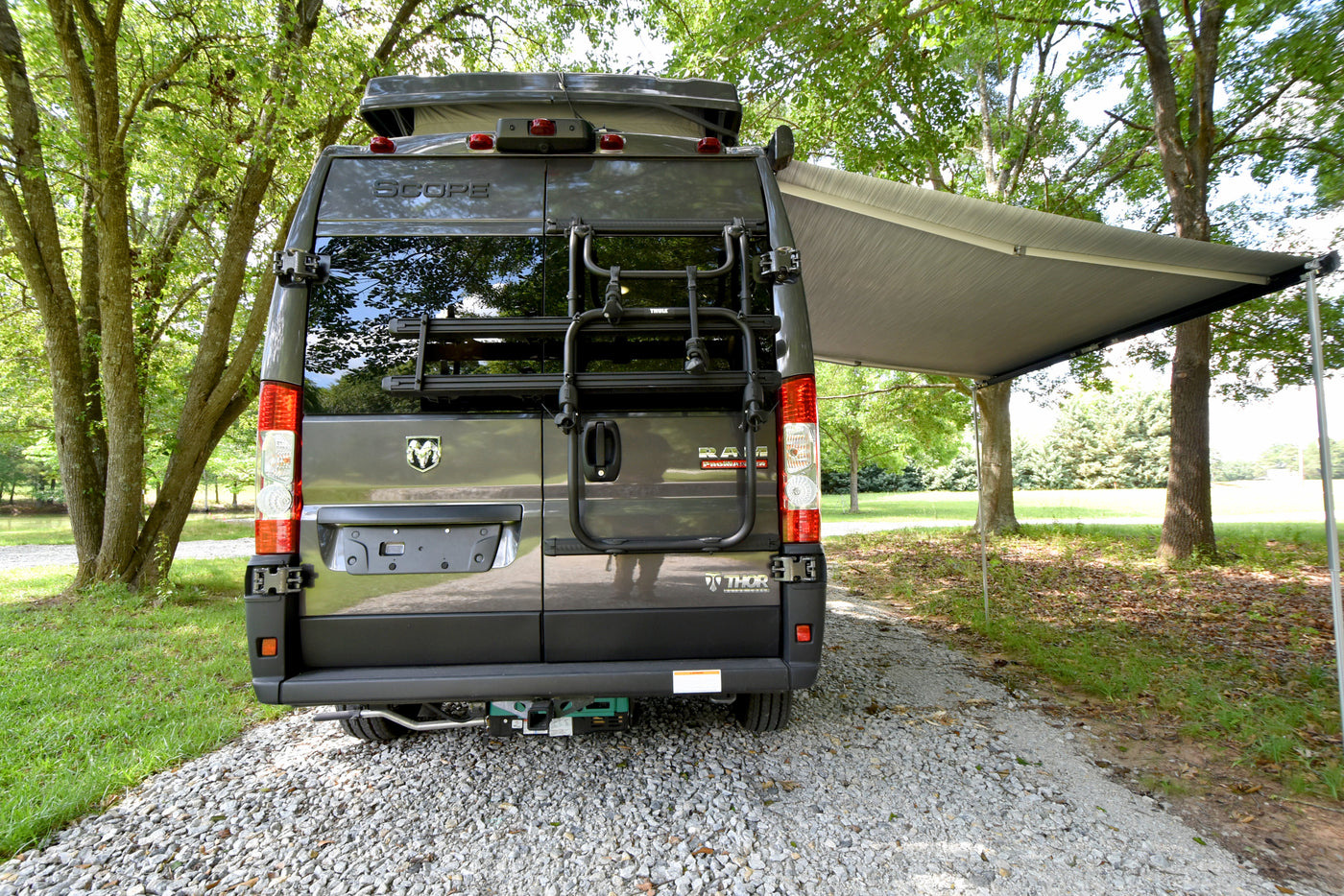 Rear of a Thor Scope camper van RV rental, featuring a bike rack and extended awning. Parked on gravel, nestled among green trees. — photo 16