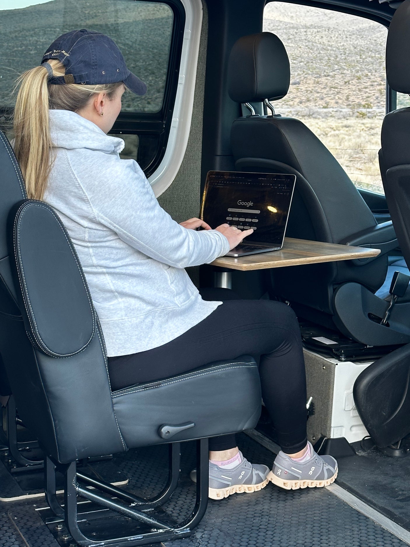 A woman in a grey hoodie & cap uses a laptop on a pull-out table inside an RV rental, viewing desert scenery.