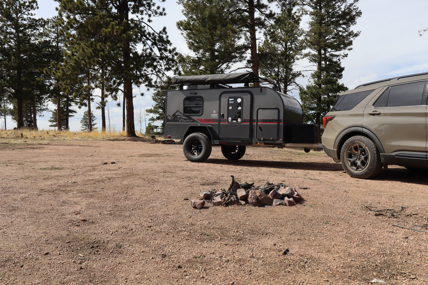A gray teardrop camper trailer towed by an SUV at a rustic campsite with a fire pit, perfect for an rv rental outdoor adventure.