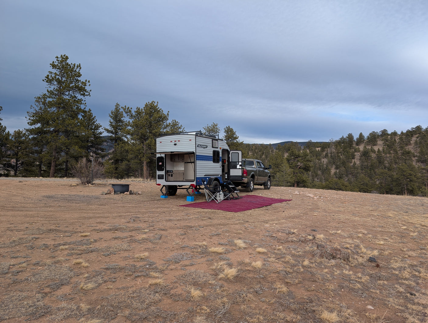 An RV rental truck camper and pickup truck parked in a scenic, dry forest clearing with a camping setup under cloudy skies.