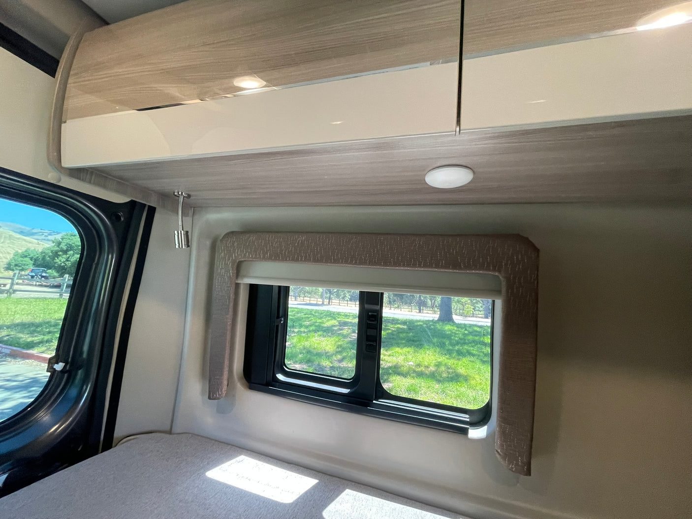 Inside an RV rental: a light-colored bed beneath a window revealing a green outdoor scene, with modern overhead cabinets and lighting.