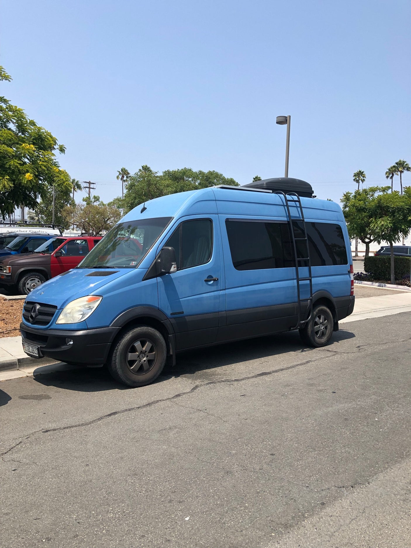 Light blue Mercedes Sprinter camper van, equipped as an RV rental with roof rack and side ladder.
