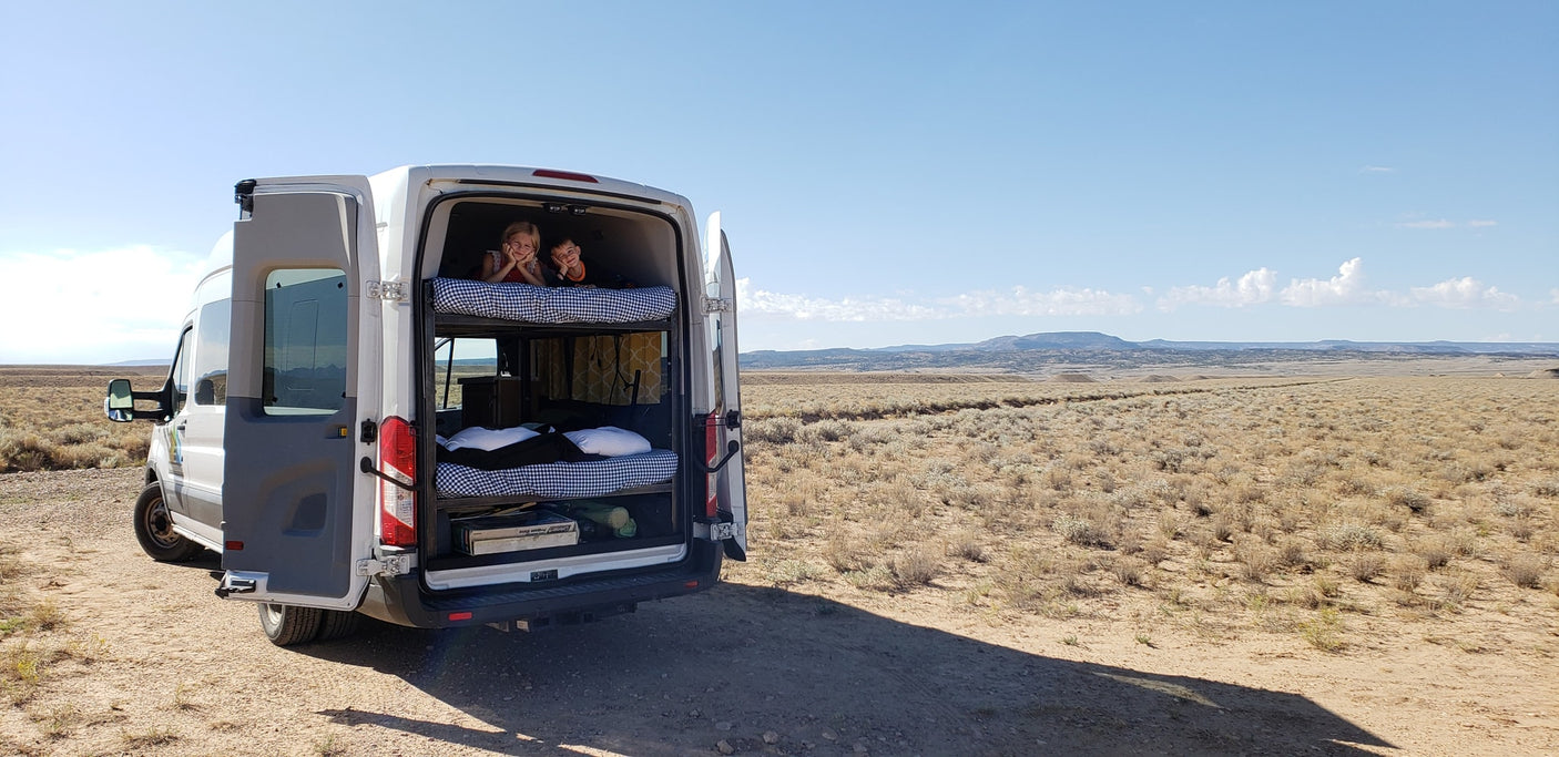 Family RV rental adventure: Kids peek from bunk beds in a campervan, parked amidst a sprawling desert landscape.
