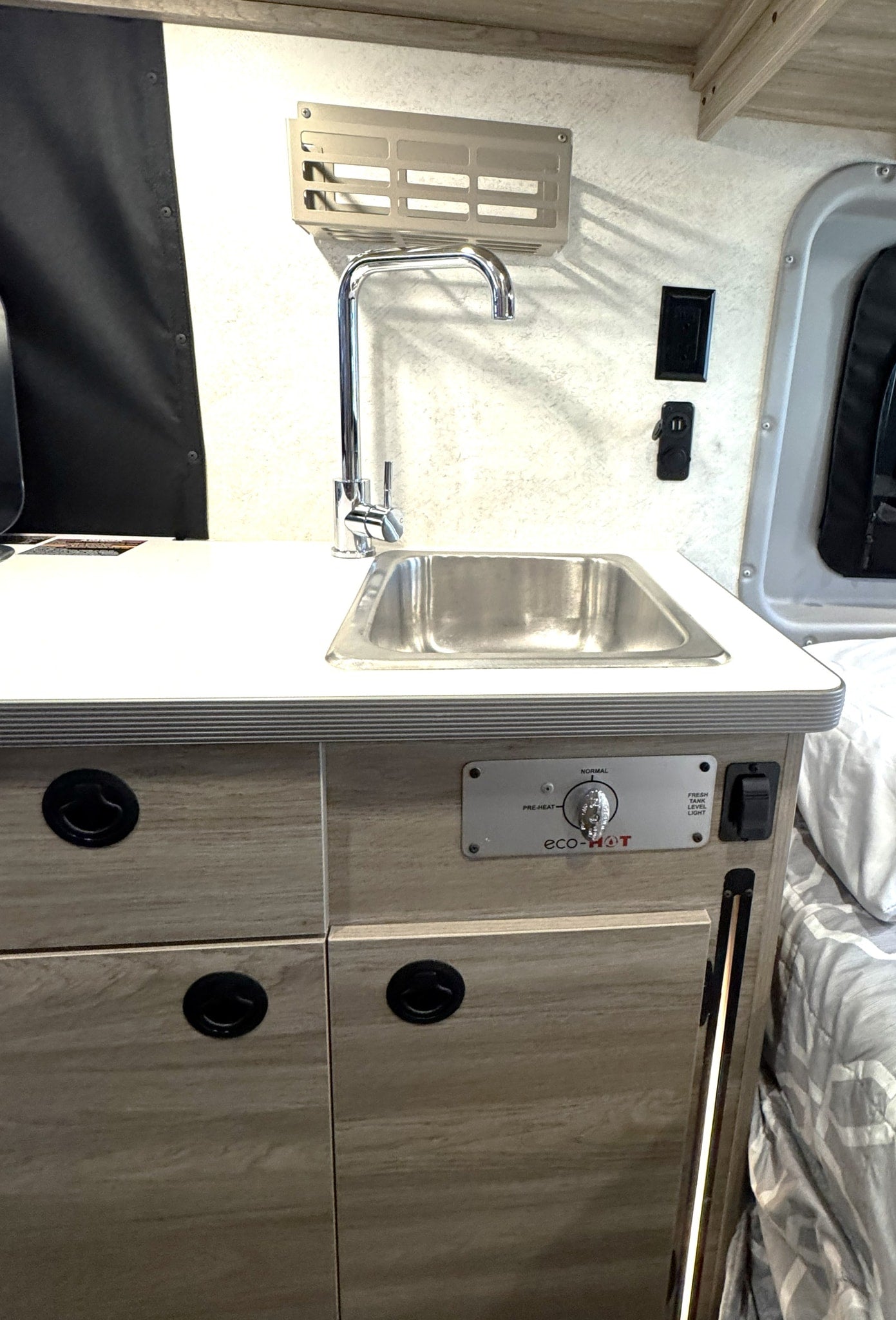 This rv rental features a compact kitchen with a stainless steel sink, chrome faucet, eco-HOT water heater controls, and light wood cabinets.