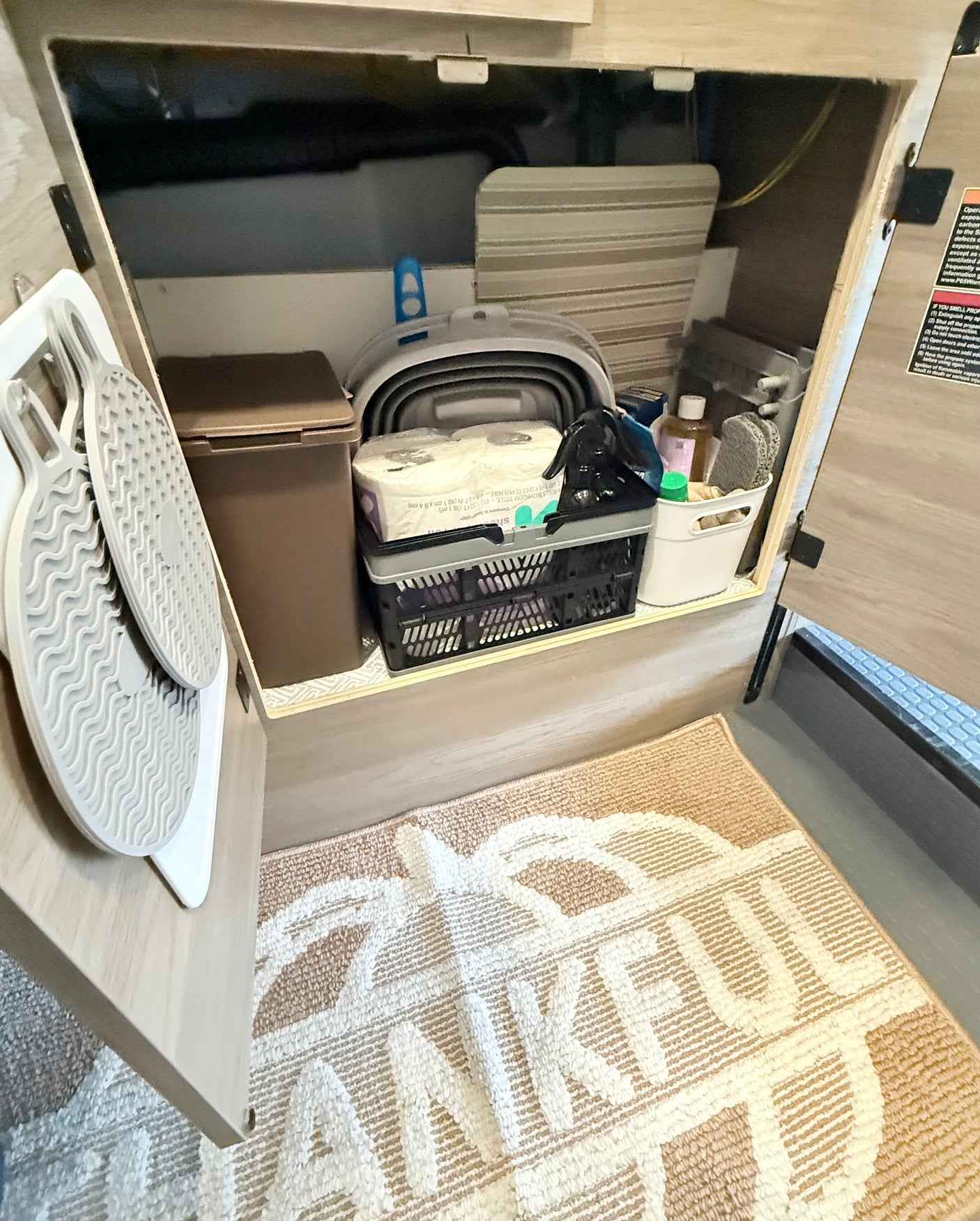 RV rental storage: cabinet with TP, cleaning supplies, and collapsible bin. Mats hang on door, "THANKFUL" rug below.
