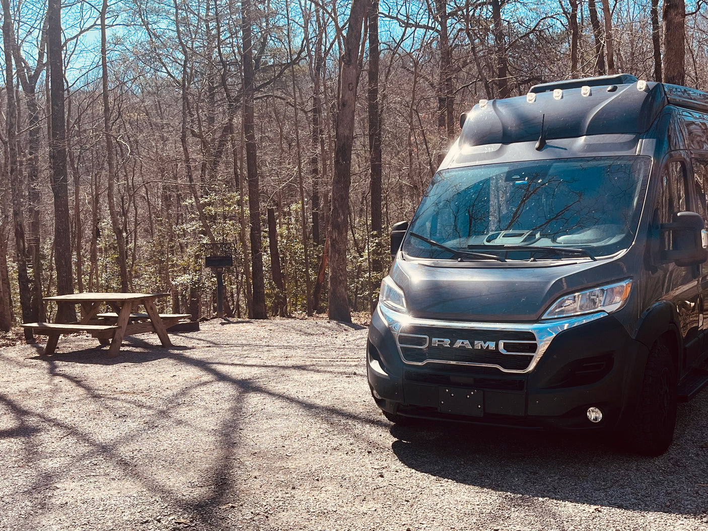 Dark grey RV rental parked on a gravel campsite with a picnic table & grill, surrounded by bare trees.