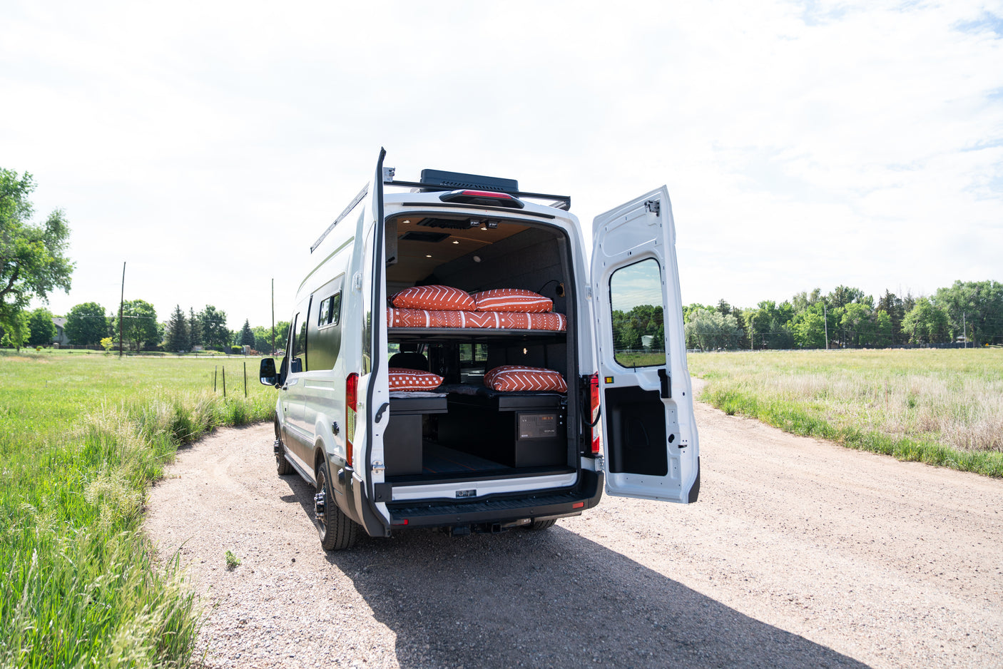 White camper van rv rental parked on a dirt road, rear doors open revealing a custom interior with beds & storage. Perfect for outdoor adventures.