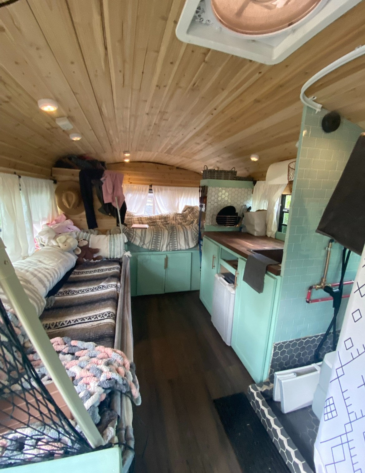 & functional kitchen space.|Pastel teal & white skoolie (converted school bus) on asphalt with open door. Unique RV rental for your next adventure!|Light blue and white Skoolie (converted school bus) at a scenic campground. Ideal for your next RV rental adventure.|Custom converted 'Skoolchella' bus in an RV campground setup with picnic gear. Explore RV rental options for travel!|Explore this cozy RV rental's interior! Wood ceilings