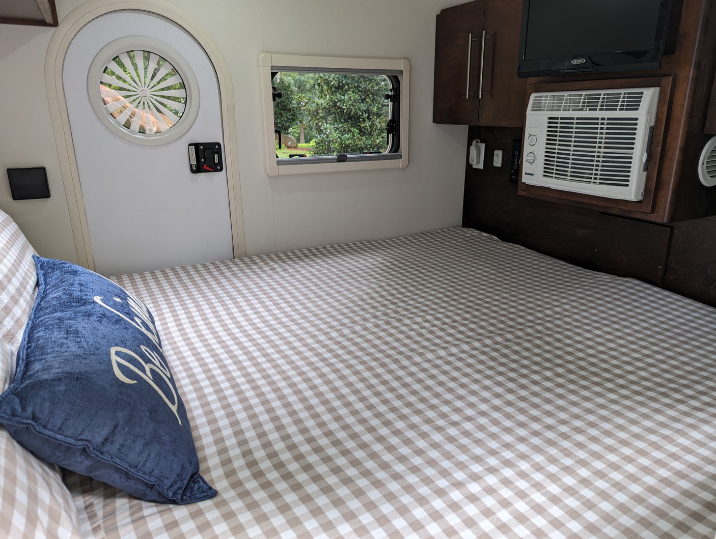 Cozy rv rental interior: checkered bed, blue pillow, outdoor window view, circular door window, AC, and TV.