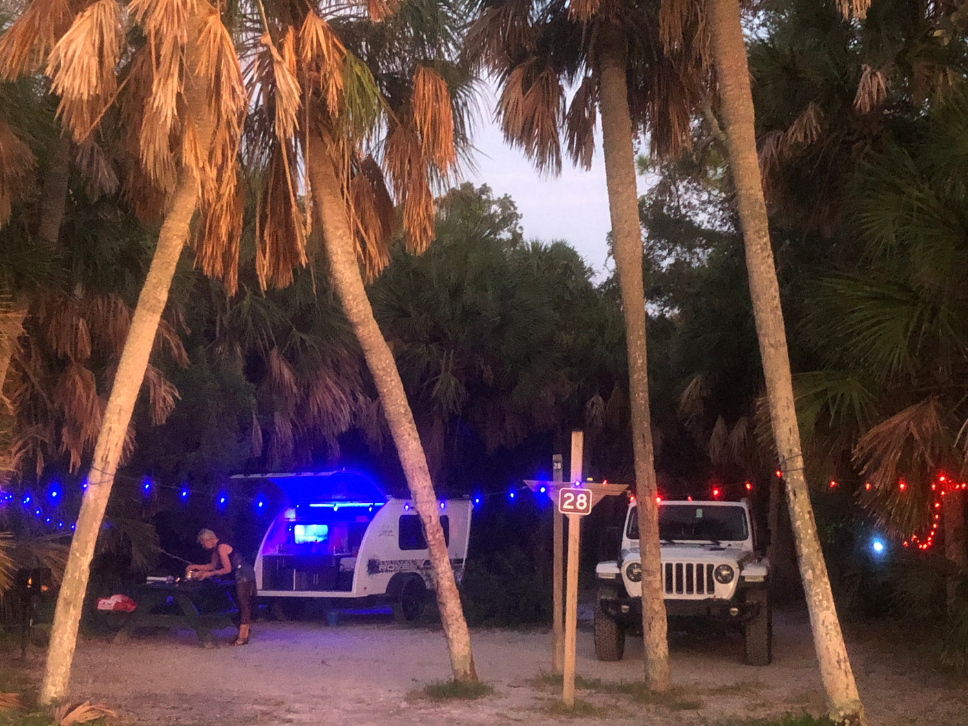 A cozy campsite with a lit teardrop RV rental, a person at a table, and a Jeep under palm trees with string lights at site 28.
