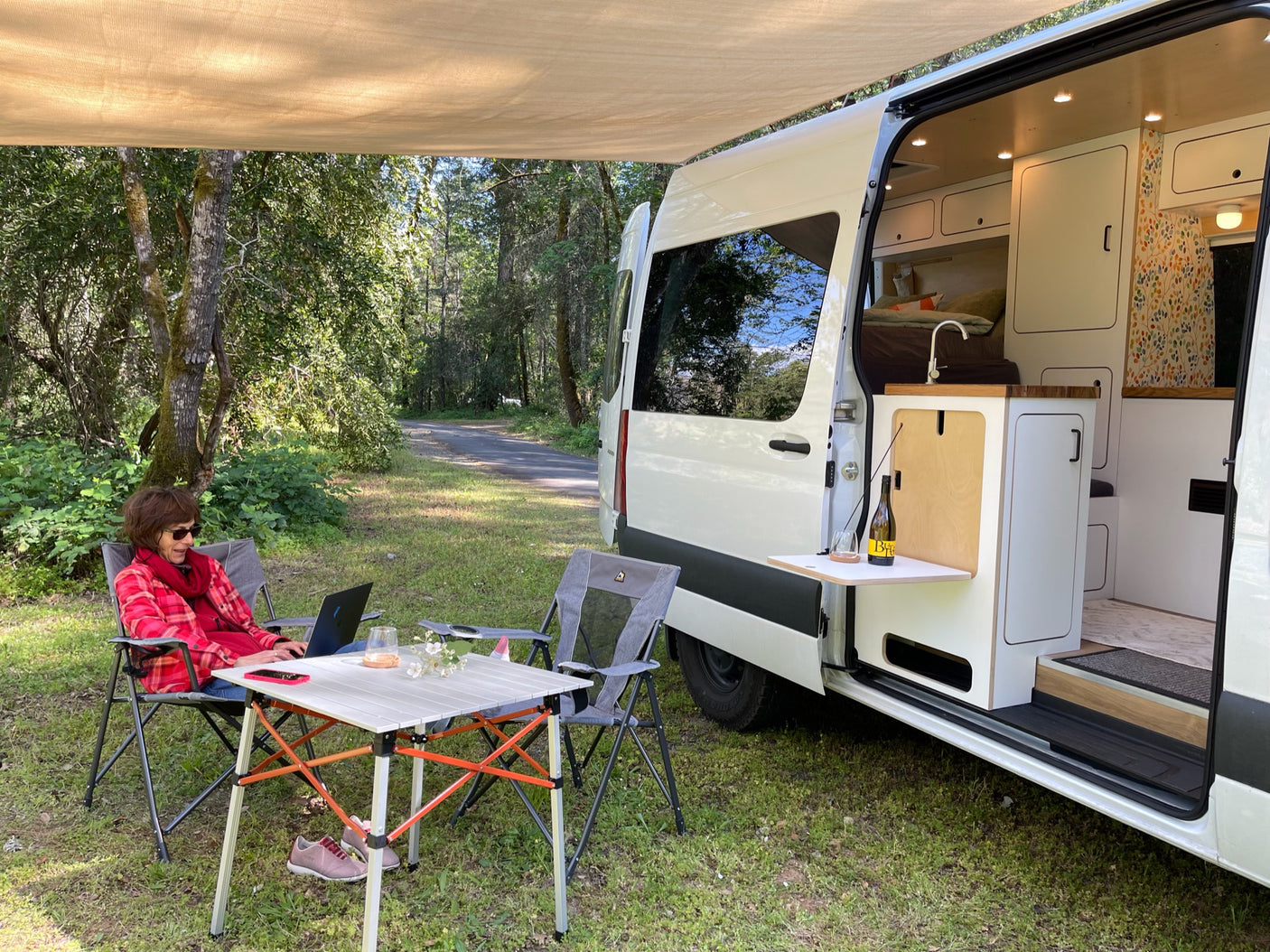 Relaxing RV rental setup: woman on laptop in red shirt, white van with open door & awning in nature.