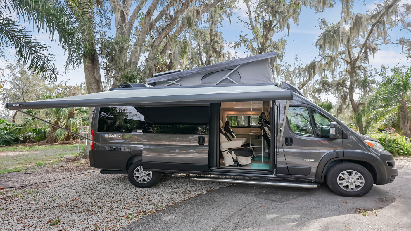Grey camper van RV rental with extended pop-top roof and awning. Parked outside, showcasing its open interior and nature backdrop.