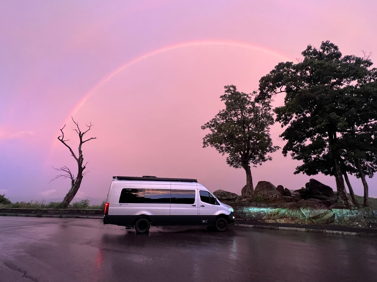 with dark trees and misty mountains.|Rear view of an Airstream Interstate X 4WD RV rental parked at a serene campground site under an awning.|Relaxing at an RV rental site with Caymus Cabernet Sauvignon wine & insulated tumblers on a picnic table.|White RV rental camper van set up at a forest campsite at dusk. Awning lights illuminate the outdoor living space.|This Mercedes-Benz Sprinter camper van — photo 5