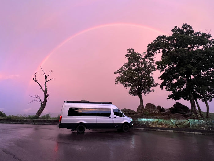 with dark trees and misty mountains.|Rear view of an Airstream Interstate X 4WD RV rental parked at a serene campground site under an awning.|Relaxing at an RV rental site with Caymus Cabernet Sauvignon wine & insulated tumblers on a picnic table.|White RV rental camper van set up at a forest campsite at dusk. Awning lights illuminate the outdoor living space.|This Mercedes-Benz Sprinter camper van