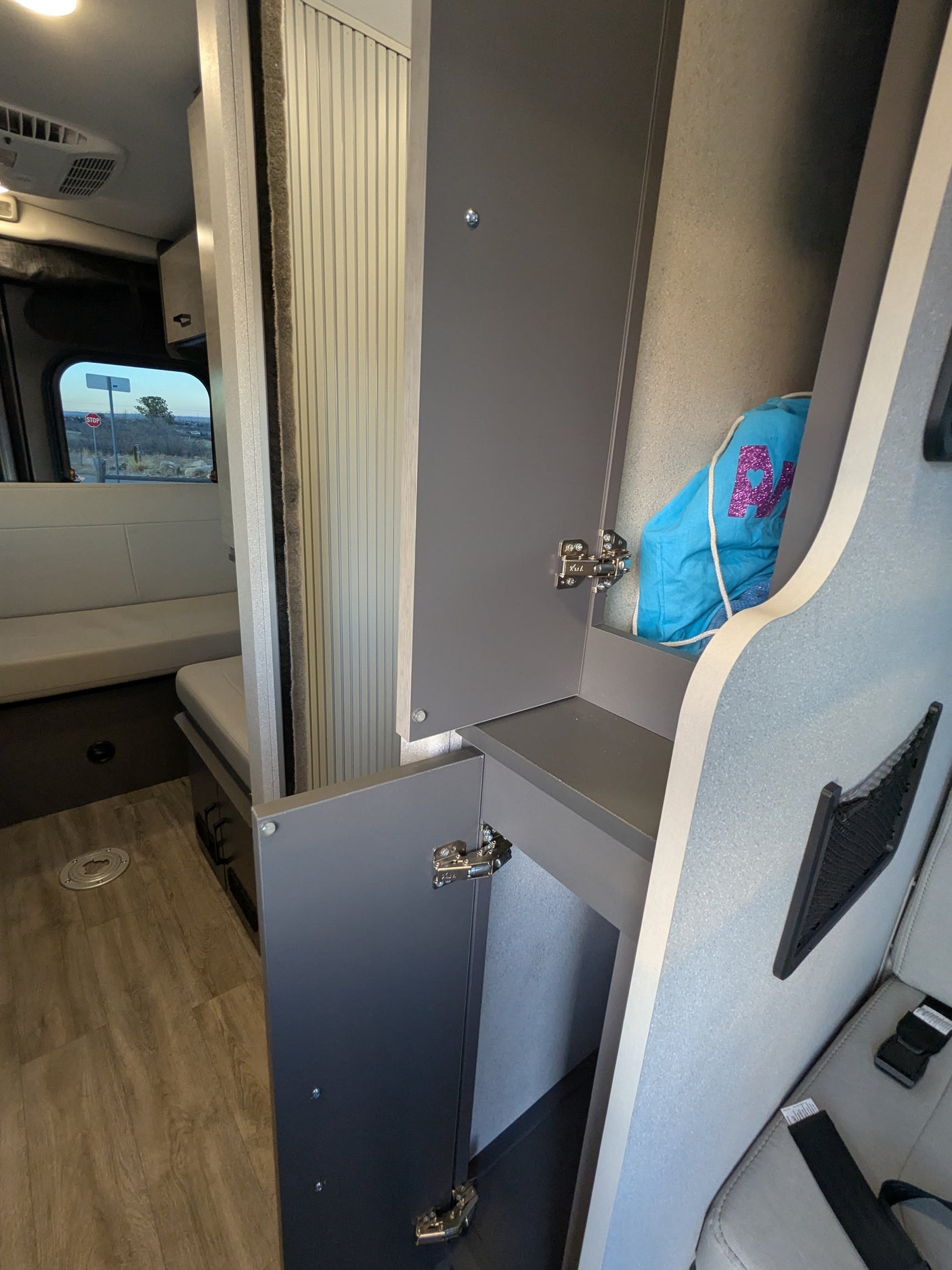 RV rental interior: open gray cabinets, light bench seating, and wood-look flooring with a window view to a stop sign.