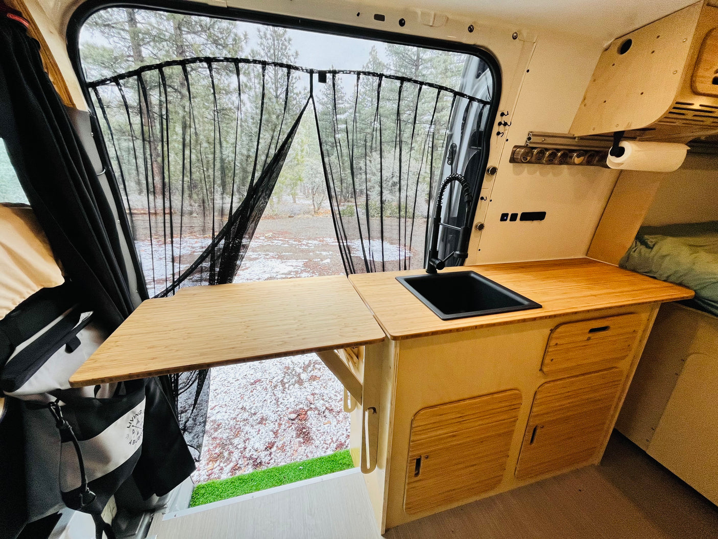 Cozy RV rental interior: kitchenette with bamboo countertops, sink, and snowy forest view through a black mesh screen.