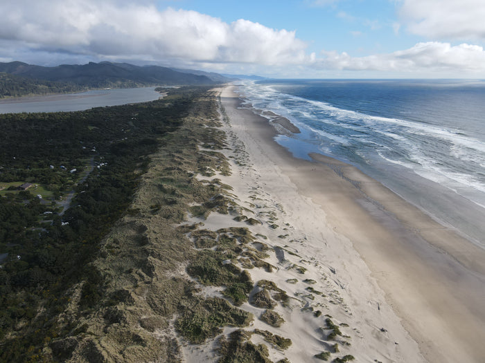 Aerial view: expansive sandy beach meeting ocean waves, backed by dunes, forest, river, and an RV rental campground.