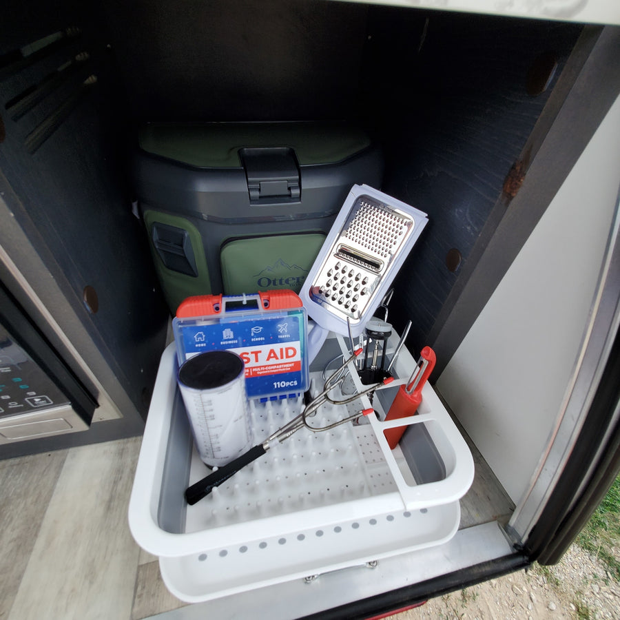 RV rental storage showing kitchen supplies (grater, measuring cup), First Aid kit, and Otter Box cooler for organized travel.