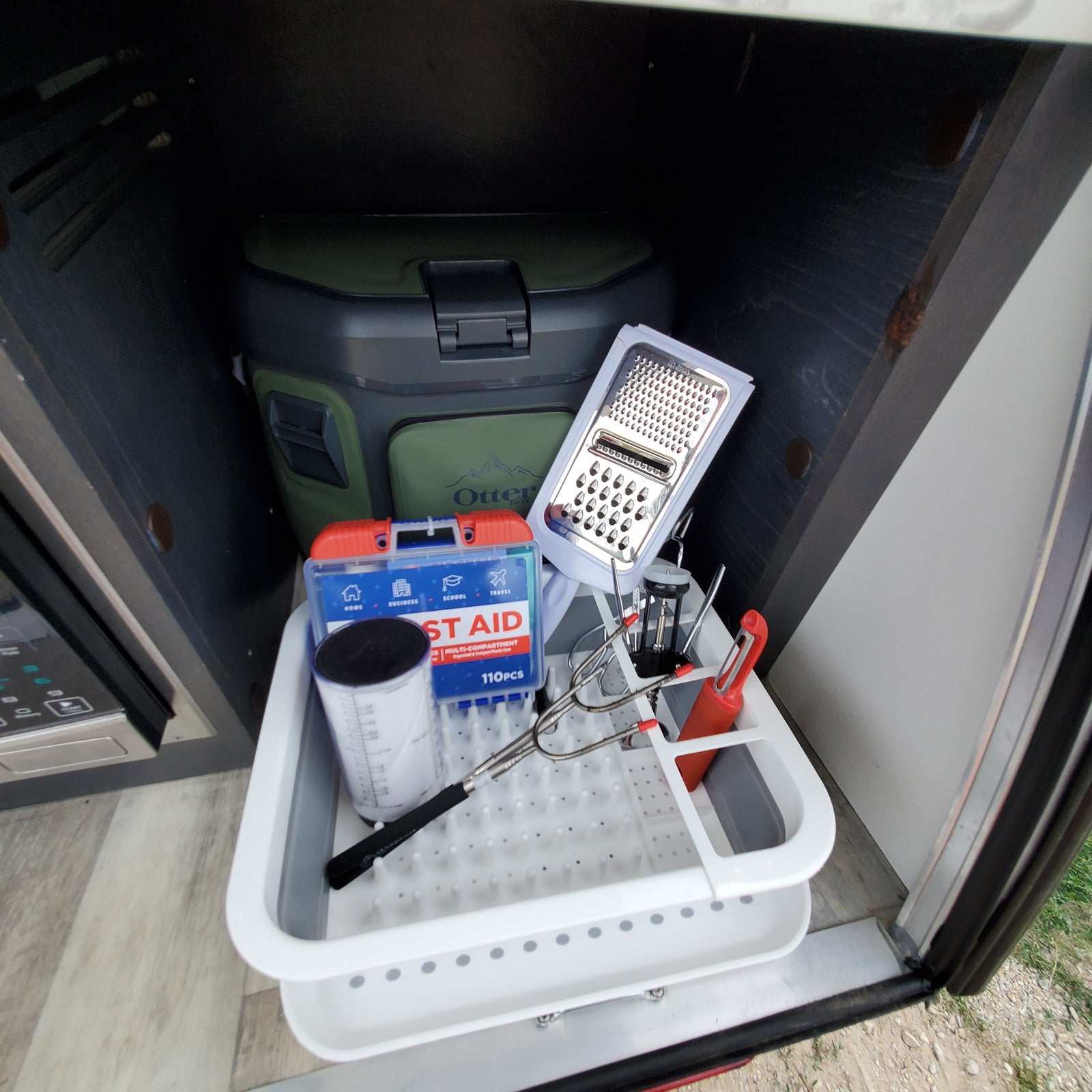 RV rental storage showing kitchen supplies (grater, measuring cup), First Aid kit, and Otter Box cooler for organized travel.