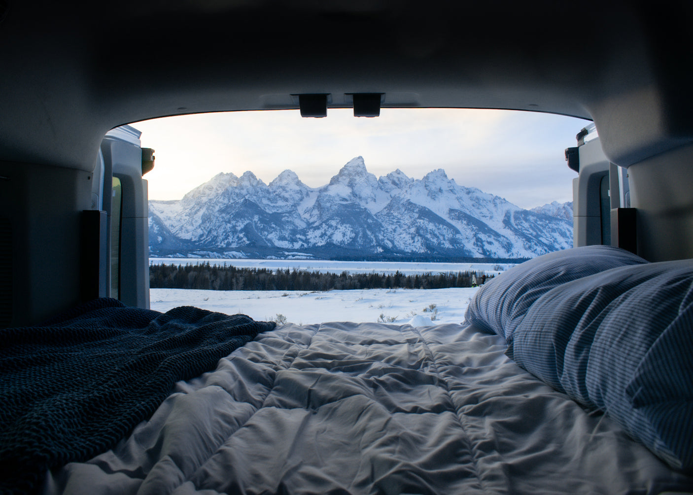 Inside an RV rental, a comfy bed overlooks a serene snow-covered landscape & majestic Teton mountains. Ultimate winter camp view.