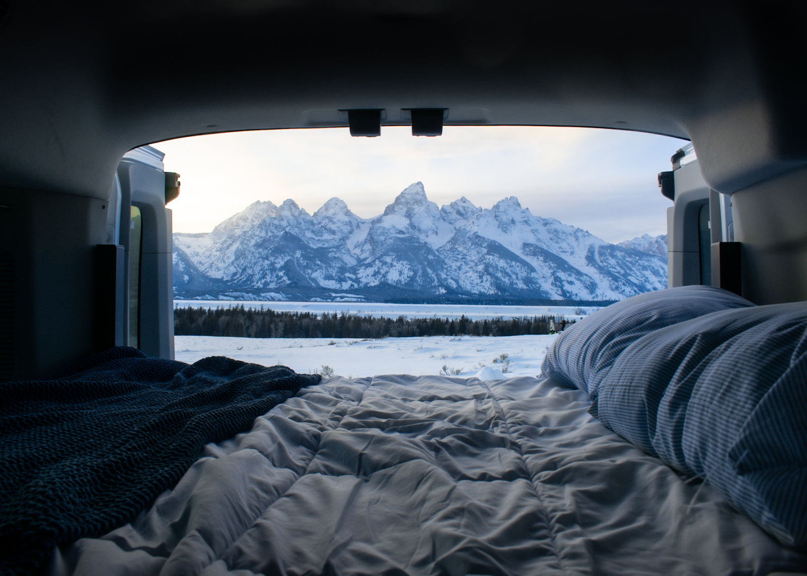 Inside an RV rental, a comfy bed overlooks a serene snow-covered landscape & majestic Teton mountains. Ultimate winter camp view.