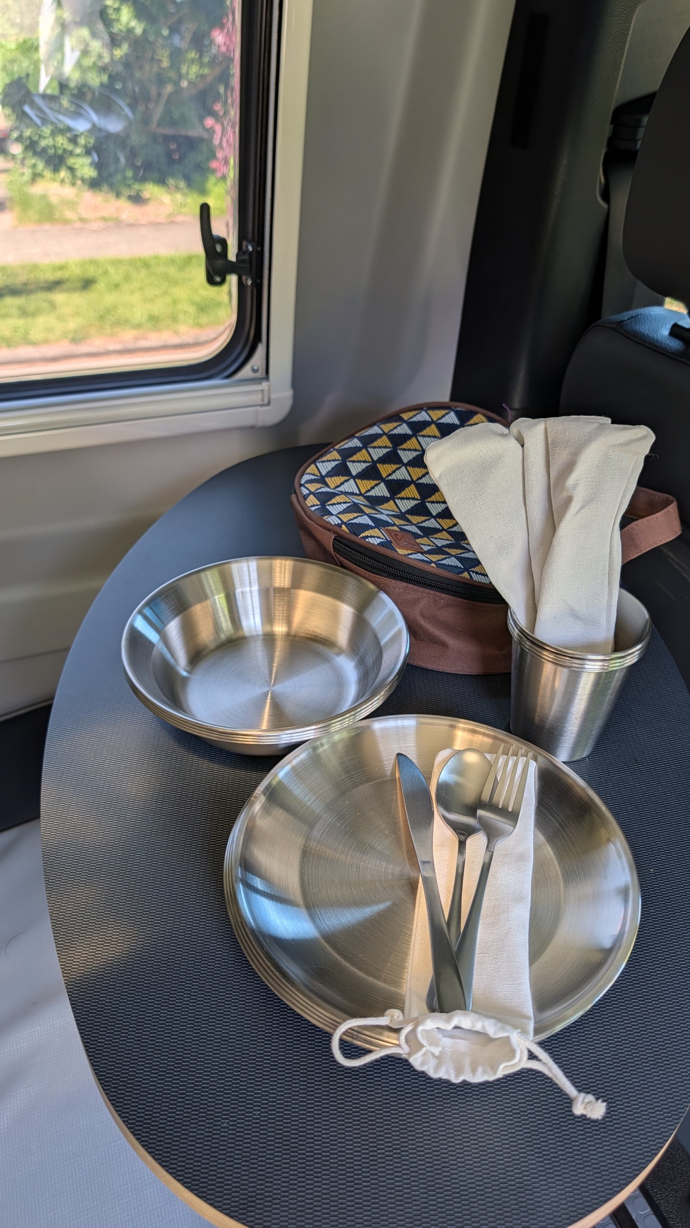 Enjoy dining in this RV rental! Stainless steel plates, cutlery, napkins & picnic bag on a textured table by a window.