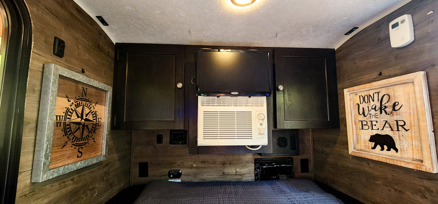 Inside a compact rv rental: wood walls, built-in TV, GE AC unit, and storage cabinets over a bed with rustic decor.