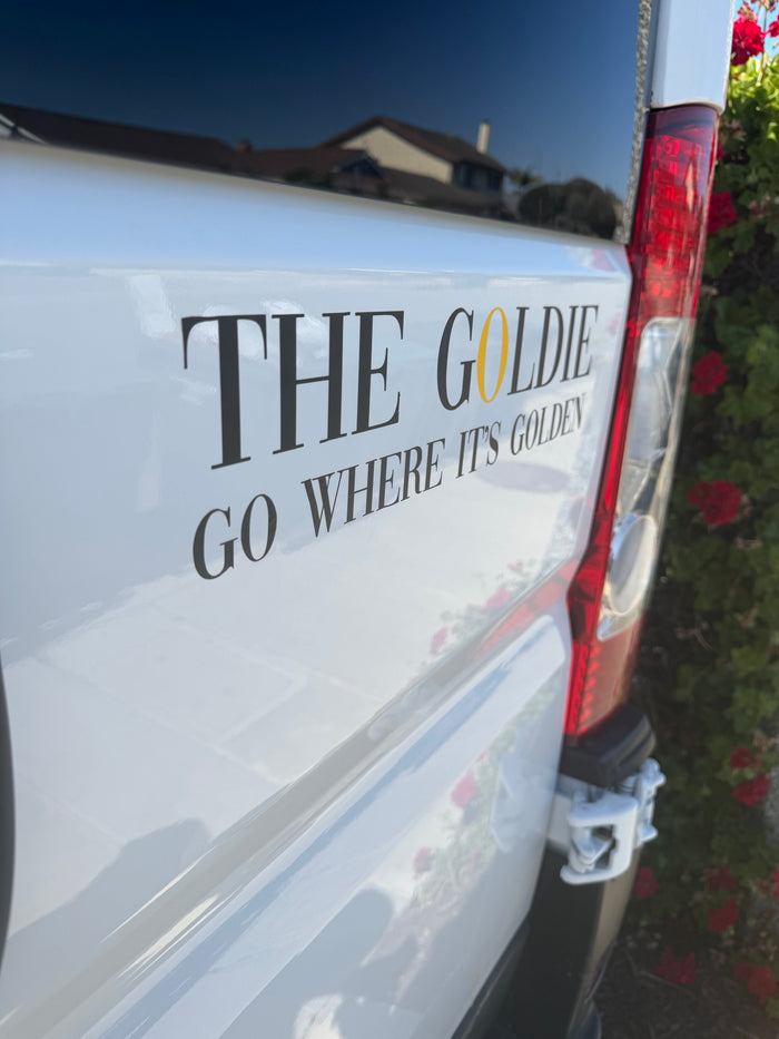 White RV rental van features "THE GOLDIE" branding with "GO WHERE IT'S GOLDEN" slogan. House reflection, red flowers visible.