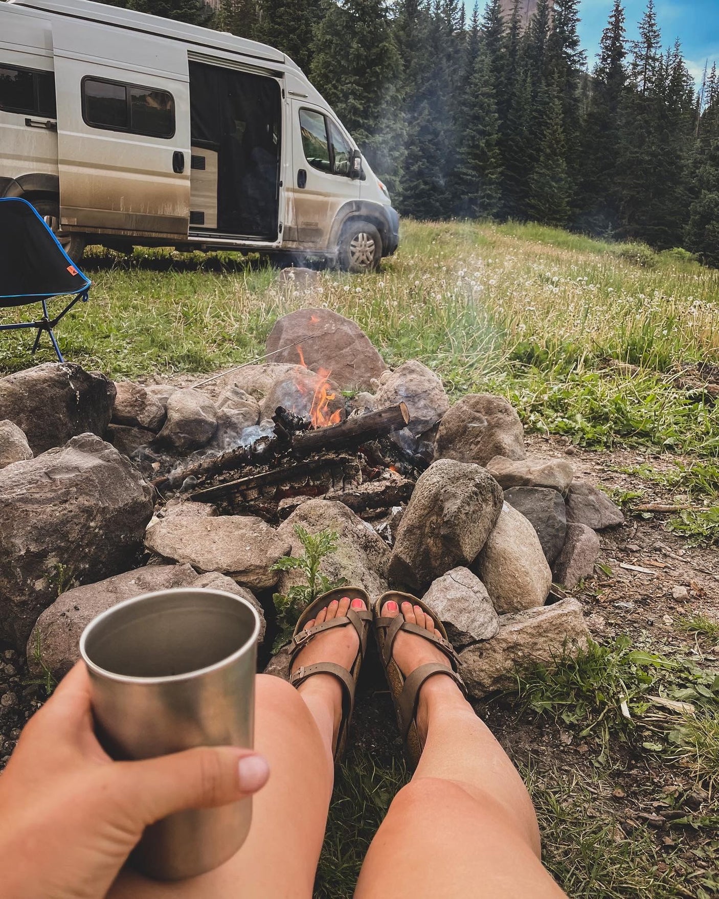 Enjoying a campfire from a ground-level view, feet propped up, with a dirty white camper van (RV rental) and trees in the background.