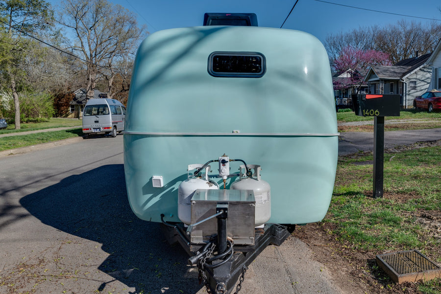 Rounded light blue camper rental with propane tanks on a residential street. A van and mailbox 1406 are visible.