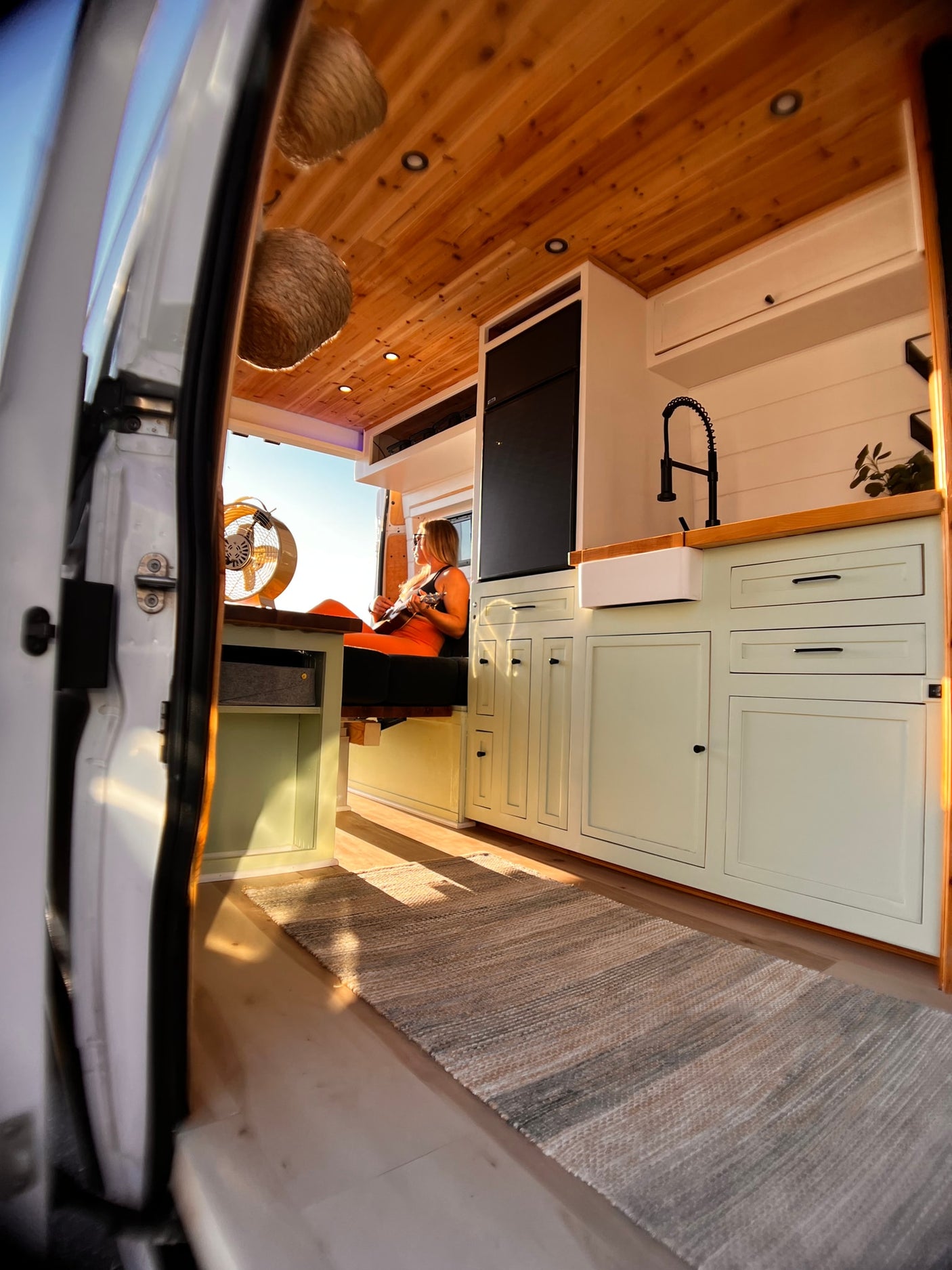 Cozy RV rental van interior. Woman plays ukulele in custom-built space with kitchen, wood ceiling, and natural light.