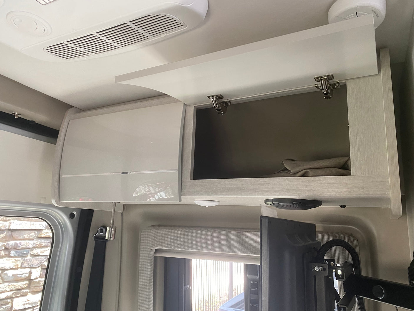 RV rental interior showcasing overhead storage cabinets (one open), ceiling AC, TV, and window with blind. Compact and organized.