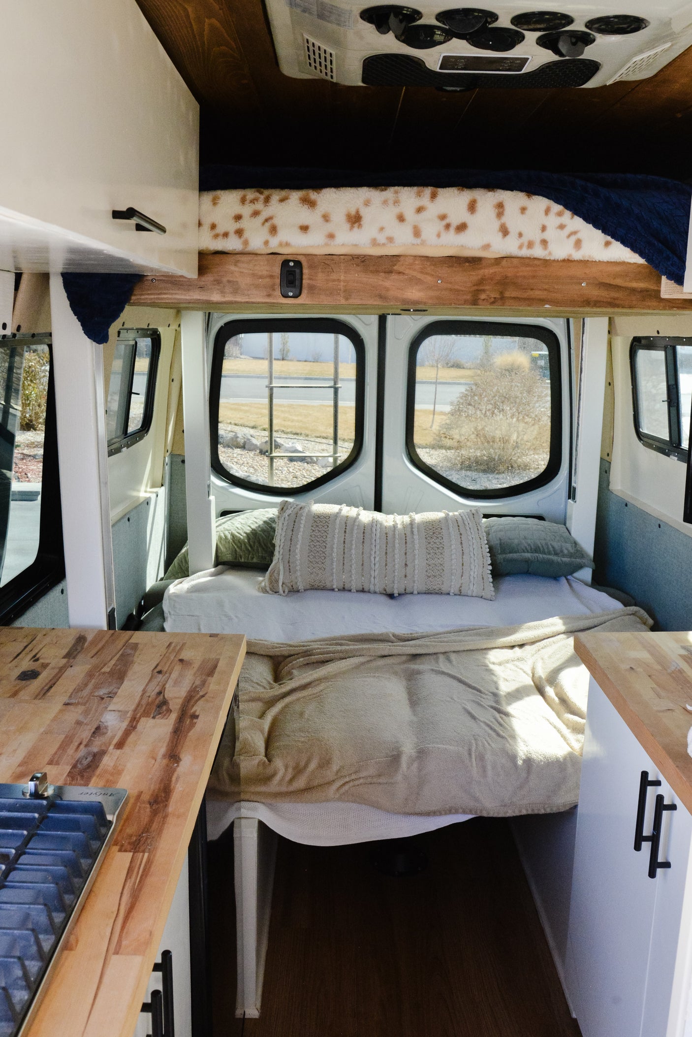 Inside a modern RV rental: a comfy bed with pillows, large windows overlooking the outdoors, and wooden kitchen counters.