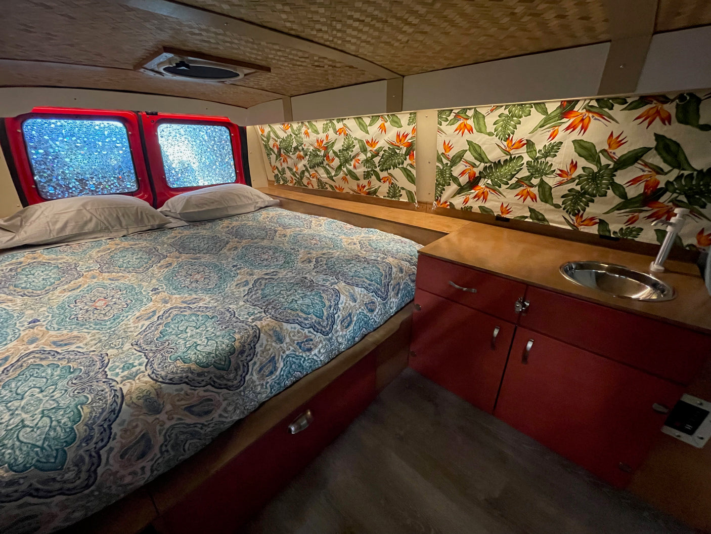 RV rental interior: bed, vibrant tropical curtains, red cabinets, sink. Woven ceiling & blurred windows add charm.