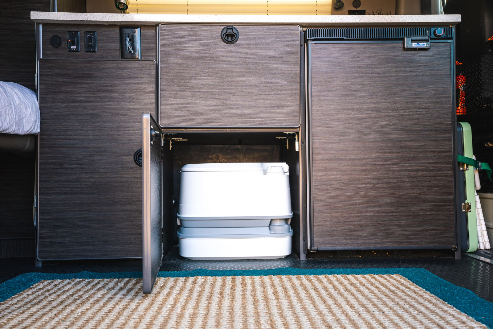 This RV rental interior shows a hidden portable toilet in cabinetry, along with a compact fridge and electrical panel, ideal for travel.