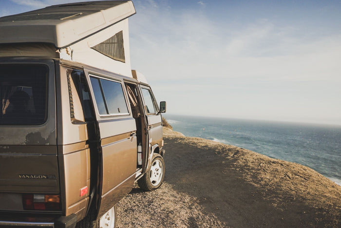 Brown VW Vanagon pop-top camper van on a coastal cliff overlooking the ocean. Perfect RV rental for your next adventure!