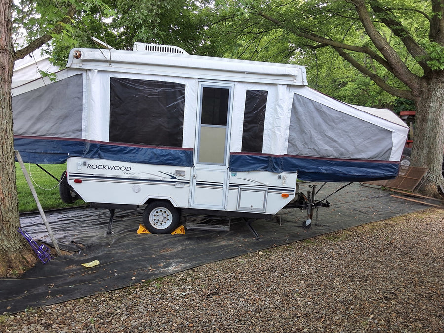 A white Rockwood pop-up camper, perfect as an RV rental or travel trailer, is set up on a black tarp among trees.