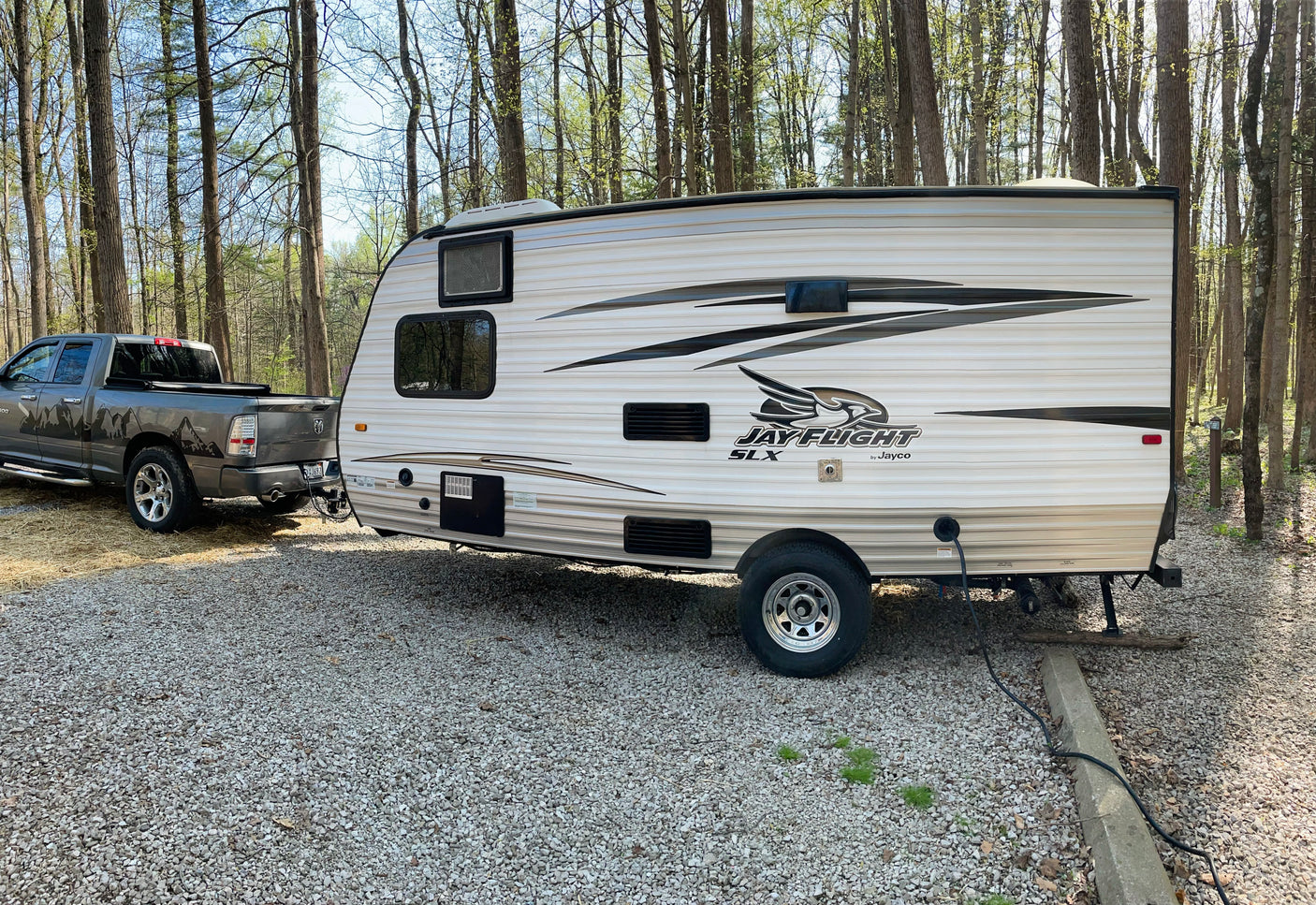 RV rental setup: A Jayco travel trailer hitched to a pickup truck on a gravel campsite amidst trees, perfect for camping.