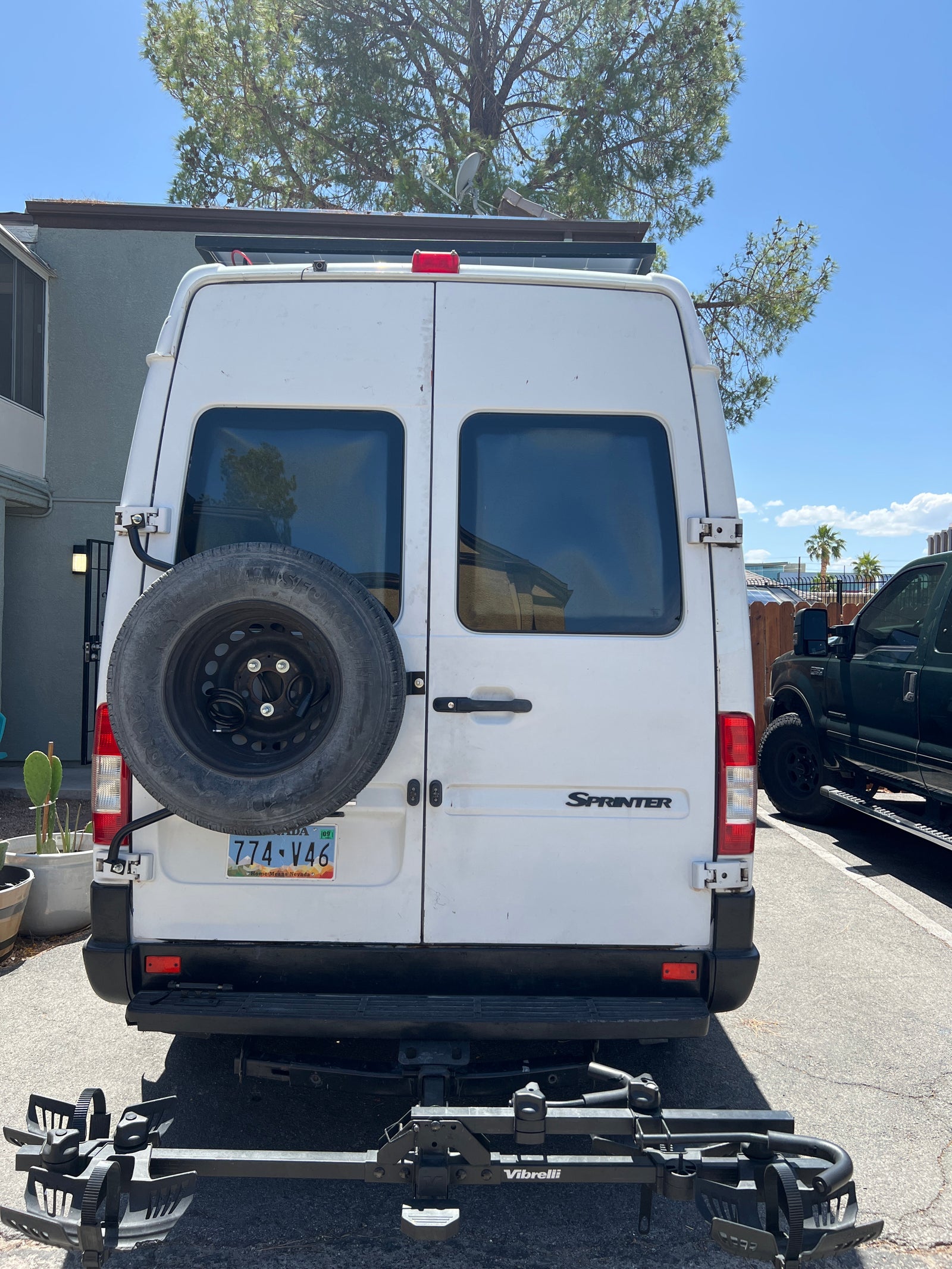 Rear view of a white Mercedes Sprinter van, a common RV rental. It has a spare tire, bike rack, and roof-mounted solar panels.