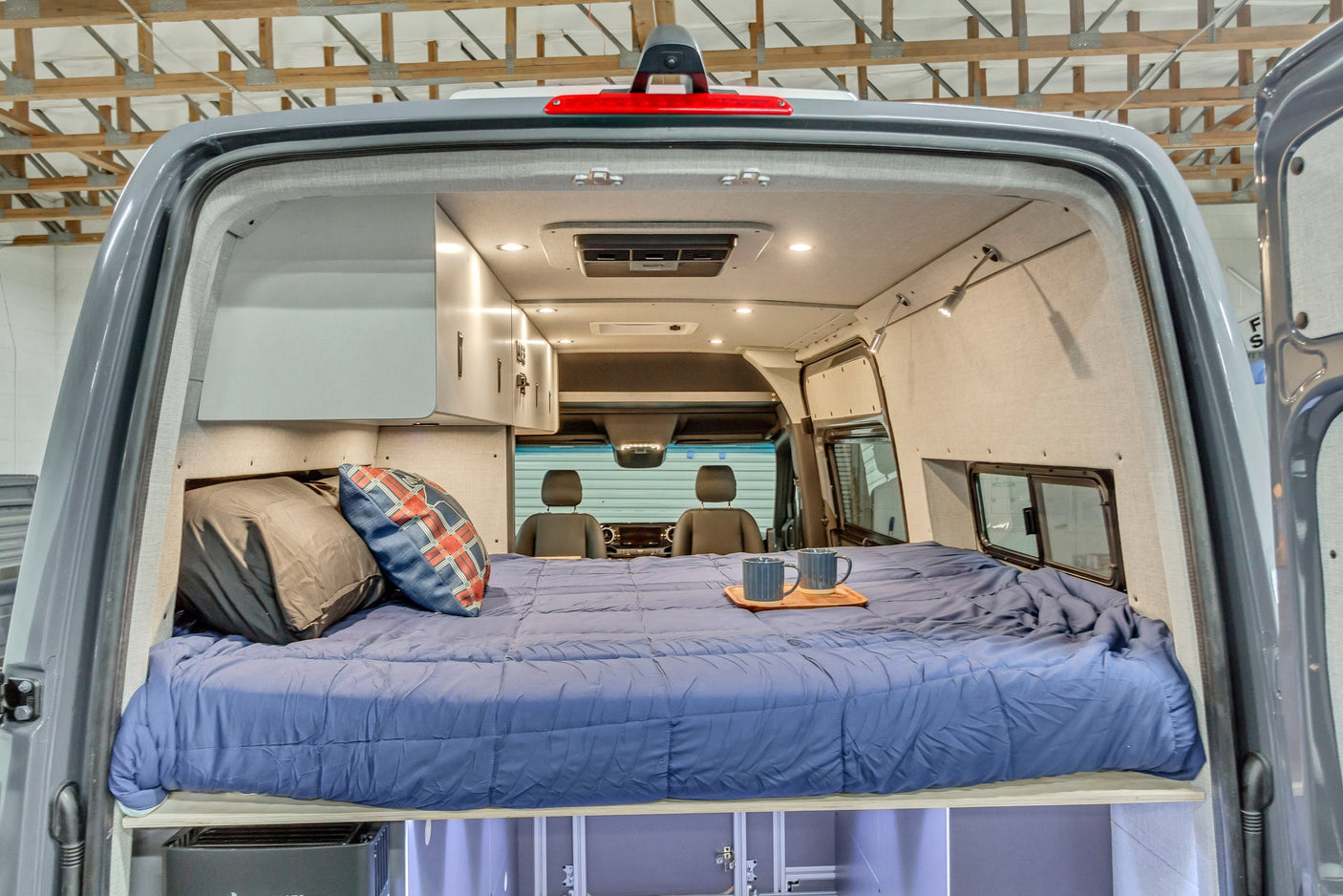 Inside a custom RV rental van: a comfy bed with blue bedding, pillows, overhead storage, and a view to the front cabin.