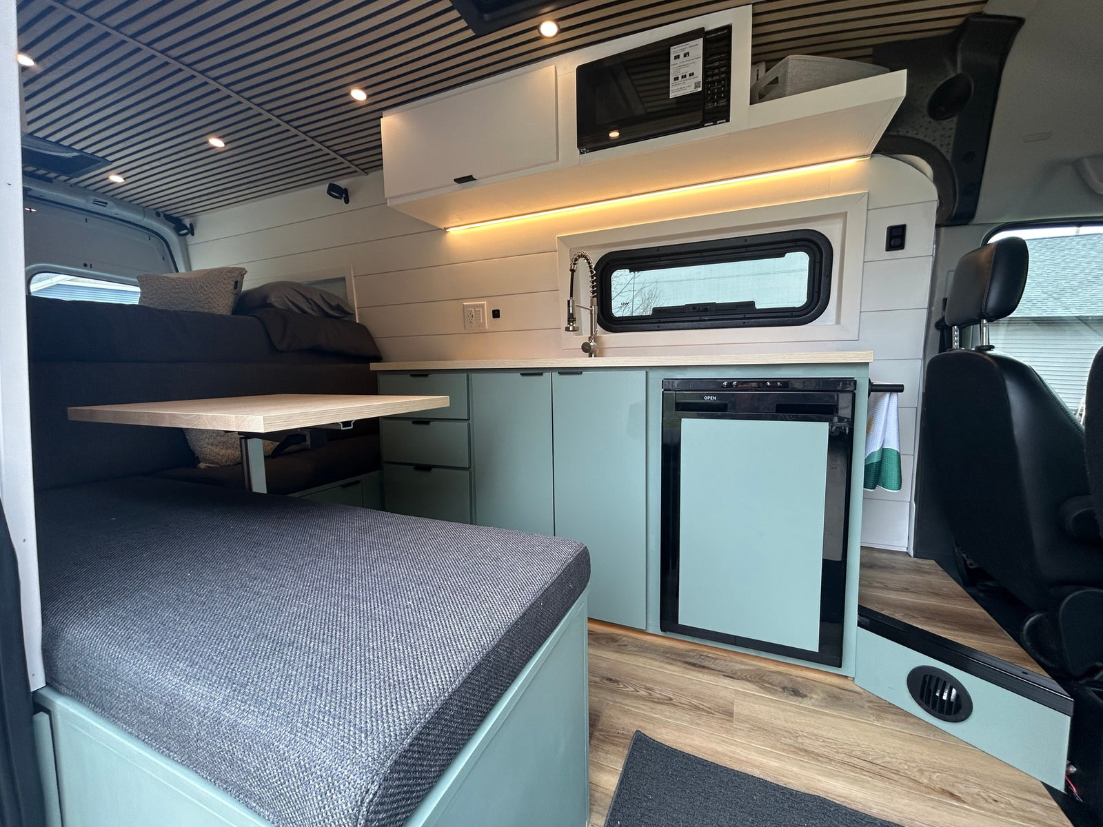 Modern RV rental interior features a mint green kitchenette with a sink, microwave, and fridge, adjacent to a bed and dining area.